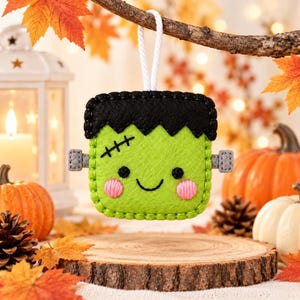 May include: A square felt ornament shaped like a green monster with black hair, a stitched scar on the forehead, two small grey bolts on the sides, black eyes, and pink cheeks, hanging from a white cord against a festive autumn background.