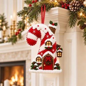 May include: A felt candy cane shaped ornament featuring a red and white striped design with white snow accents. The ornament includes a small house with a red door, a wreath, windows, a chimney, and a hanging lantern, suspended by a red ribbon.