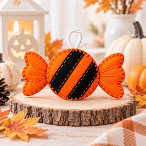May include: A felt candy-shaped ornament featuring a bright orange circular center with two diagonal black stripes and orange side wrappers. The edges are finished with visible orange stitching, and a white twisted rope loop is attached to the top.