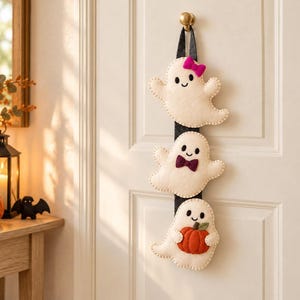 May include: A vertical wall hanging featuring three cream-colored felt ghosts attached to a black fabric strap. The top ghost wears a pink bow, the middle ghost has a dark purple bowtie, and the bottom ghost holds a small orange felt pumpkin.