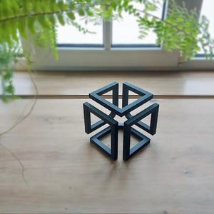 Könnte beinhalten: Eine mattschwarze geometrische Skulptur mit einem unmöglichen Würfeldesign. Das Objekt besteht aus miteinander verbundenen quadratischen Rahmen, die eine durchgehende, nicht-euklidische Form mit scharfen Kanten und einem hohlen Zentrum bilden, platziert auf einer flachen Oberfläche.