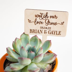 May include: A small succulent plant in an orange pot with a wooden sign that reads "watch our love grow 05.04.22 BRIAN & GAYLE"