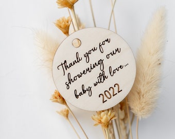 May include: A round wooden tag with the text "Thank you for showering our baby with love... 2022" engraved on it. The tag is surrounded by dried flowers.