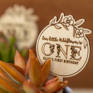 May include: A wooden sign with a floral design and the text "Our little Wildflower is ONE Ellie's First Birthday".