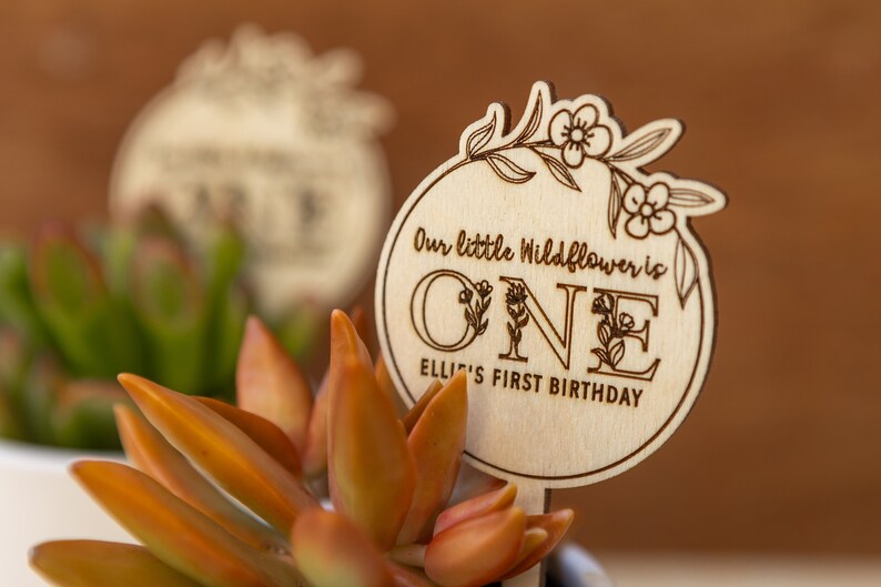 May include: A wooden sign with a floral design and the text "Our little Wildflower is ONE Ellie's First Birthday".