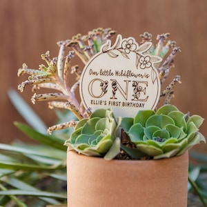 May include: A wooden sign with the text "Our little Wildflower is ONE Ellie's First Birthday" in a circle with floral designs. The sign is placed in a pot with a succulent plant.
