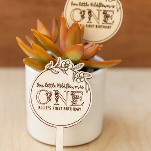May include: Wooden sign with a floral design and the text "Our little Wildflower is ONE Ellie's First Birthday".