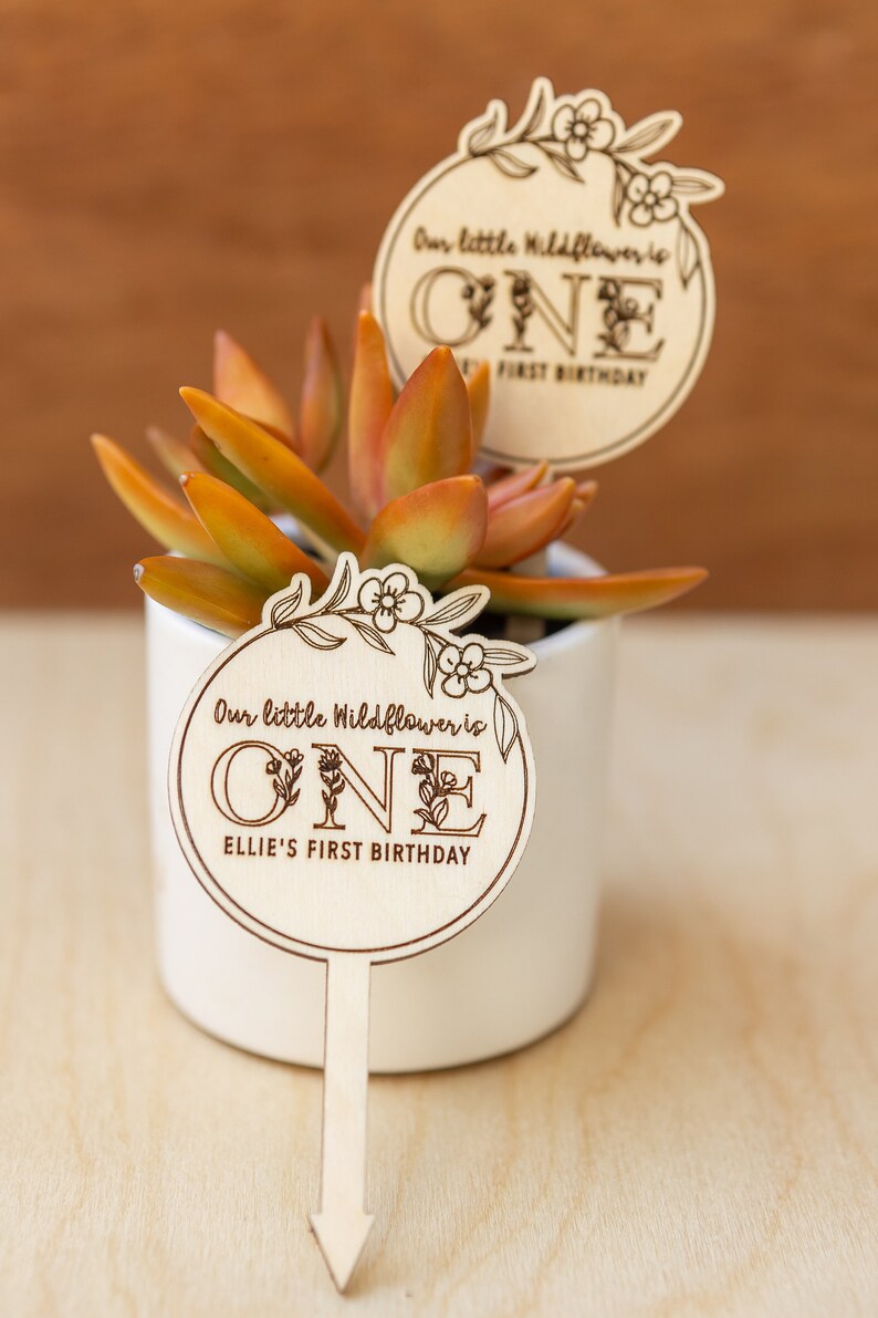 May include: Wooden sign with a floral design and the text "Our little Wildflower is ONE Ellie's First Birthday".