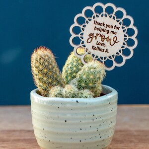 May include: A small cactus plant in a white ceramic pot with a wooden sign that says "Thank you for helping me grow, Love, Kollins A."