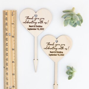 May include: Two wooden heart-shaped wedding favor signs with a stick on the back. The signs say "Thank you for celebrating with us!" with the names "Nezir & Christina" and the date "September 10, 2022".