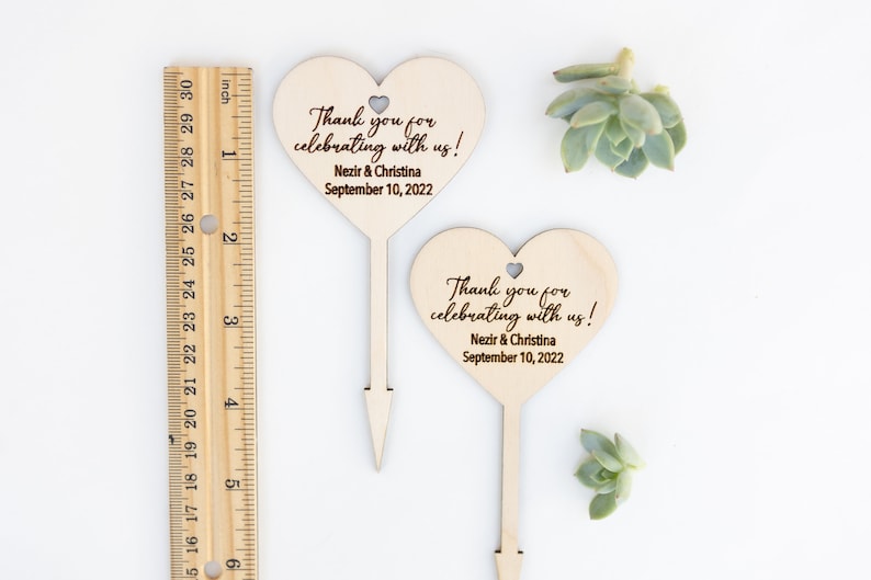 May include: Two wooden heart-shaped wedding favor signs with a stick on the back. The signs say "Thank you for celebrating with us!" with the names "Nezir & Christina" and the date "September 10, 2022".