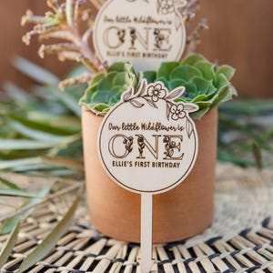 May include: Wooden sign with a floral design and the text "Our little Wildflower is ONE Ellie's First Birthday". The sign is attached to a wooden stick and is placed in a pot with a succulent plant.