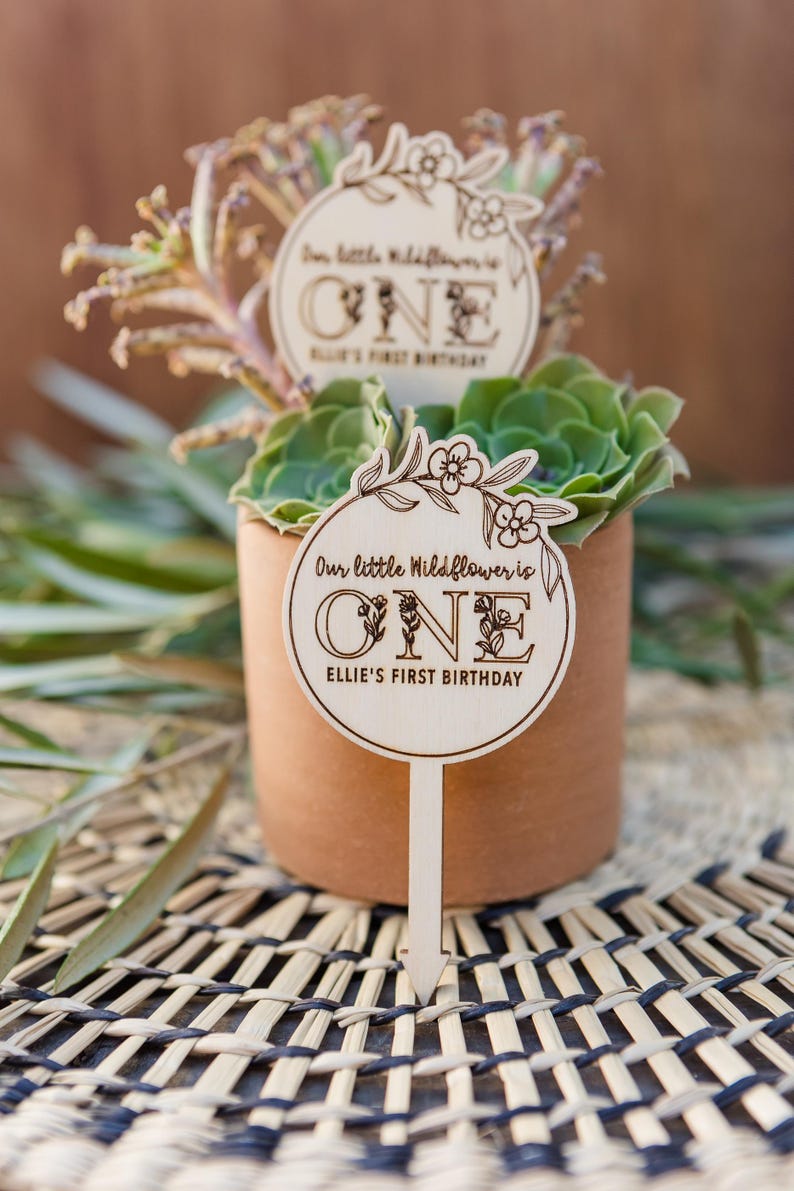 May include: Wooden sign with a floral design and the text "Our little Wildflower is ONE Ellie's First Birthday". The sign is attached to a wooden stick and is placed in a pot with a succulent plant.
