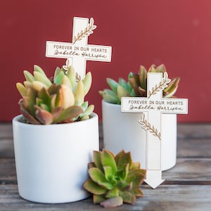 May include: Two white wooden crosses with the text "Forever in our hearts" and a name on each. The crosses are placed in small white pots with green succulents.