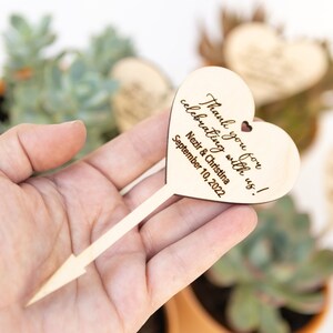 May include: Wooden heart-shaped sign with a stick attached. The sign says "Thank you for celebrating with us!  Nezir & Christina September 10, 2022".