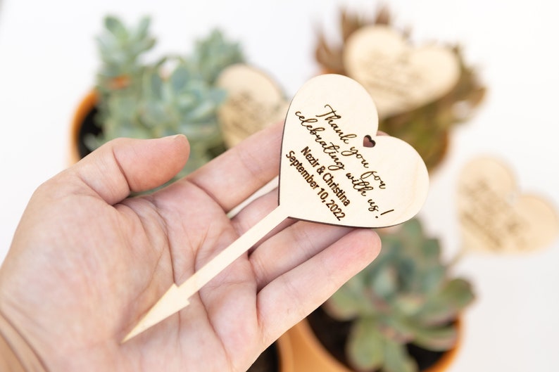 May include: Wooden heart-shaped sign with a stick attached. The sign says "Thank you for celebrating with us!  Nezir & Christina September 10, 2022".
