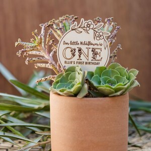 May include: A wooden sign with the words "Our little Wildflower is ONE Ellie's First Birthday" and floral designs. The sign is propped up in a terracotta pot with two green succulents.