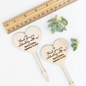 May include: Two wooden heart-shaped wedding favor signs with the text "Thank you for celebrating with us! Nezir & Christina September 10, 2022".