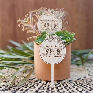 May include: A wooden sign with a floral design and the text "Our little wildflower is ONE Ellie's First Birthday" is stuck into a small potted succulent plant.