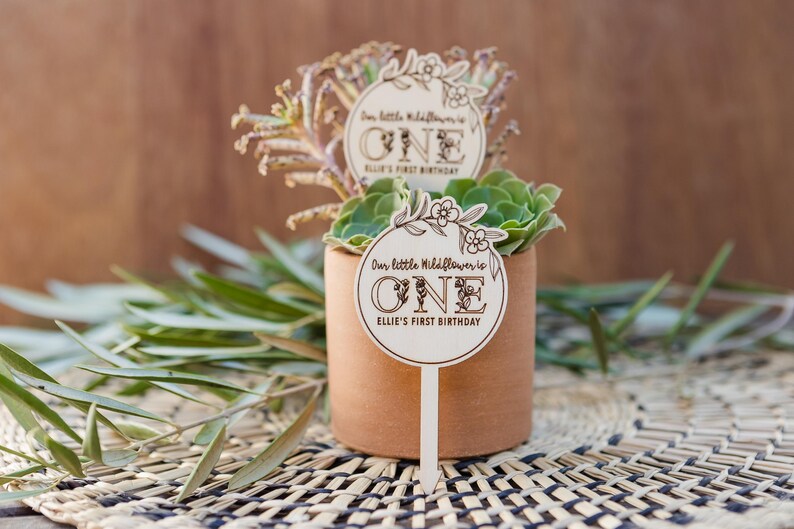 May include: A wooden sign with a floral design and the text "Our little wildflower is ONE Ellie's First Birthday" is stuck into a small potted succulent plant.