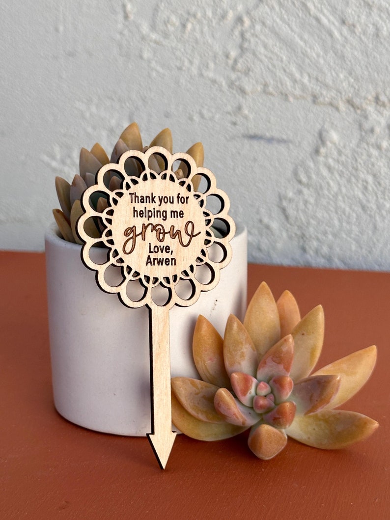 May include: Wooden plant marker with a decorative edge and the text "Thank you for helping me grow, Love, Arwen".