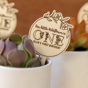May include: A wooden sign with the text "Our little wildflower is one Ellie's first birthday" with floral designs around the number one. The sign is attached to a small potted succulent.