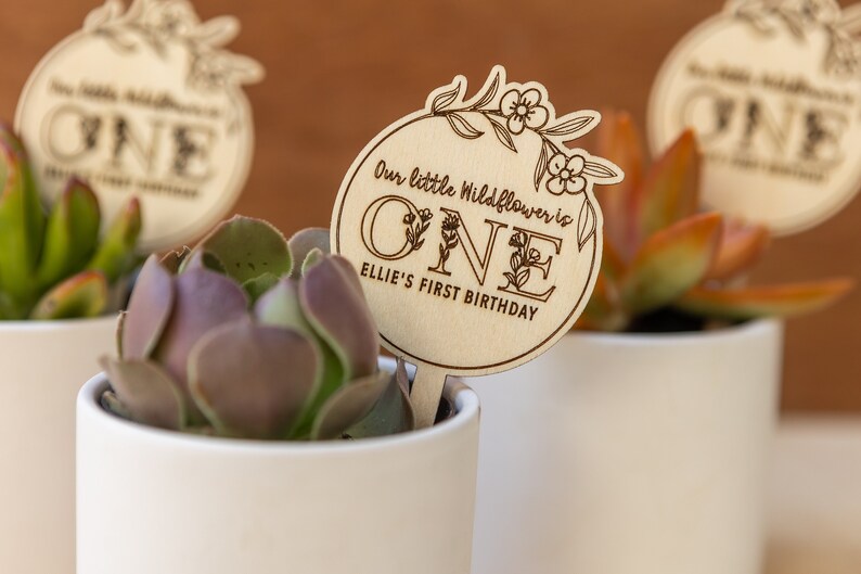 May include: A wooden sign with the text "Our little wildflower is one Ellie's first birthday" with floral designs around the number one. The sign is attached to a small potted succulent.