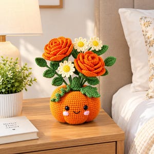 Könnte beinhalten: Ein gehäkelter orangefarbener Kürbis mit lächelndem Gesicht, rosa Wangen und schwarzen Augen, der als Basis für einen Blumenstrauß aus zwei orangefarbenen Rosen, drei weißen Gänseblümchen mit gelber Mitte und grünen Blättern aus Garn dient.