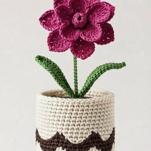 May include: A crocheted magenta flower with a green stem and two leaves, planted in a cylindrical cream-colored pot. The pot features a dark brown zigzag pattern around the middle and a single horizontal brown stripe near the base.