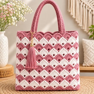 Puede incluir: Un bolso de ganchillo con un patrón de conchas repetitivo en hilo de color rosa empolvado y crema alternados. El bolso tiene dos asas a juego de color rosa y una borla decorativa de hilo rosa sujeta a una cadena dorada en la parte frontal.