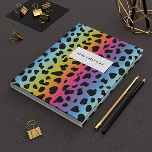 May include: A rectangular notebook featuring a vibrant rainbow gradient background with a black leopard print pattern. A white rectangular label on the cover displays the black text ADHD DAILY PLAN. The notebook is accompanied by two pencils.