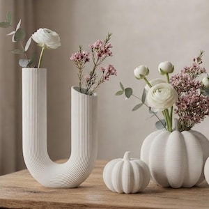 Op de afbeelding: Een witte U-vormige vaas met ribbels met een witte bloem en groene bladeren, naast een witte geribbelde pompoenvormige vaas met witte en roze bloemen, en twee extra witte geribbelde pompoenvormige decoratieve objecten op een houten oppervlak.