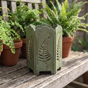 Op de afbeelding: Een saliegroene keramische plantenbak met een zeshoekige vorm en geschulpte bovenranden. Elk van de zes zijden heeft een verticaal uitgesneden ontwerp dat lijkt op een varenblad. De plantenbak staat op kleine voetjes en heeft een gladde glazuurlaag.