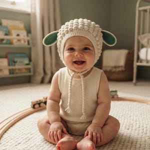 May include: A cream-colored crochet hat featuring a textured bobble stitch pattern, two protruding green and cream ears, and braided chin ties with tassels. The hat is worn over a simple, sleeveless cream-colored onesie made of soft fabric.