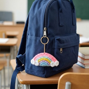 May include: A dark blue canvas backpack with a gold-toned metal keychain attached to the zip. The keychain features a small, handmade crocheted rainbow with pink, yellow, light blue, and purple stripes, finished with white clouds on each end.