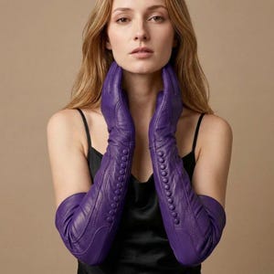 Könnte beinhalten: Ein Paar lange, ellbogenlange Handschuhe aus violettem Leder mit einer vertikalen Reihe passender, stoffbezogener Knöpfe entlang des Unterarms. Die Handschuhe weisen sichtbare Ziernähte und eine glatte, matte Oberfläche auf.