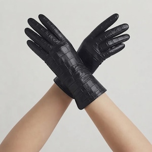 Könnte beinhalten: Ein Paar schwarze Handschuhe mit einer strukturierten Krokodilleder-Optik. Die Handschuhe sind überkreuzt angeordnet und zeigen die glänzende Oberfläche sowie das detaillierte geprägte Schuppenmuster an den Fingern und am Handgelenk.