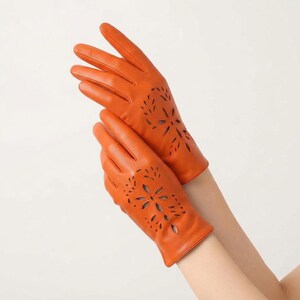 May include: A pair of vibrant orange leather gloves featuring intricate laser-cut floral patterns on the back of the hands. The gloves extend to the wrists and showcase a smooth, uniform finish with precise cutouts that form a symmetrical petal design.