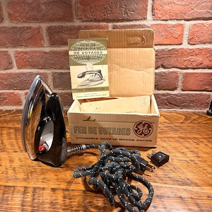 May include: A vintage General Electric travel iron with a chrome finish and black handle, accompanied by its original cardboard box, instruction manual, and a black power cord with a plug adapter. The box features the text FER DE VOYAGES.
