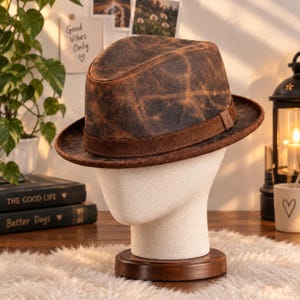 Peut inclure: Un chapeau fedora marron avec une texture vieillie et tachetée, doté d'une bande marron foncé autour de la calotte. Le chapeau est présenté sur un support de tête de mannequin de couleur neutre, mettant en valeur le bord structuré.
