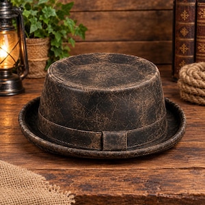 Puede incluir: Un sombrero pork pie con un acabado desgastado en marrón oscuro y negro que presenta una textura agrietada similar al cuero. El sombrero tiene una copa plana, un ala estrecha y una banda a juego alrededor de la base con un detalle rectangular.