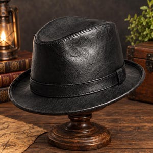Könnte beinhalten: Ein schwarzer Fedora-Hut mit strukturierter Oberfläche und einem passenden schwarzen Band um die Krone. Der Hut verfügt über eine eingekniffene Oberseite und eine strukturierte Krempe, die ein durchgehend dunkles Finish aufweist.