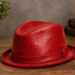 May include: A vibrant red fedora hat featuring a smooth, textured finish and a matching red band encircling the base of the crown. The hat has a structured shape with a pinched top and a wide, flat brim, showcasing a consistent deep red colour.