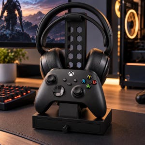 May include: A black gaming headset stand with a hexagonal cutout pattern, featuring a black wireless game controller mounted on the front and a small storage drawer at the base. The controller has a white Xbox logo and colored A, B, X, Y buttons.