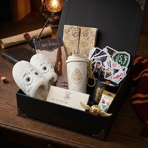 Puede incluir: Una caja de regalo negra que contiene artículos temáticos de Harry Potter: pantuflas de peluche blancas de búho, una taza de viaje con un escudo dorado, una vela negra, una snitch dorada, el Mapa del Merodeador, una carta para el Sr. H. Potter y pegatinas.