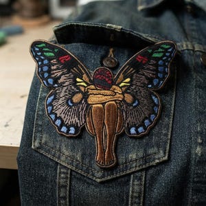 May include: An embroidered patch featuring a figure with brown skin curled into a fetal position, adorned with large, detailed butterfly wings in shades of black, blue, green, and red. The patch is attached to the pocket of a dark blue denim jacket.