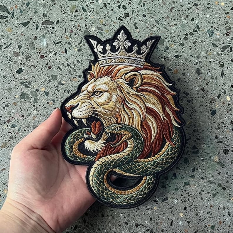 May include: An embroidered patch featuring a lion with a golden mane wearing a silver crown, with a green snake coiled around the lion's neck. The design is outlined in black thread and features detailed stitching in shades of gold, brown, and green.