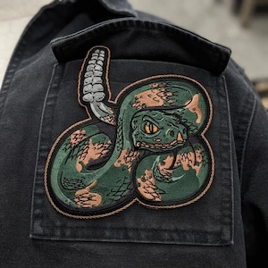 May include: An embroidered patch featuring a coiled rattlesnake with green and copper-coloured scales, a raised rattle, and a sharp, focused eye. The patch is stitched onto a black fabric garment, showcasing detailed thread work and a dark border.