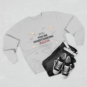 May include: A light grey crewneck sweatshirt featuring the black and red text POV: YOU'RE OVERTHINKING AGAIN, surrounded by small thought bubbles with phrases like WHAT IF, WHY, and DID I SAY THAT, paired with dark grey jeans and black trainers.