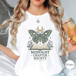May include: A white short-sleeve t-shirt featuring a graphic print of a moth above an open book, with the text "MIDNIGHT CHAPTER SOCIETY" below. The design includes a crescent moon, stars, and floral elements in shades of green, gold, and black.